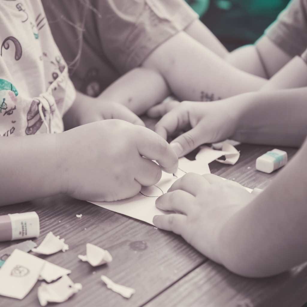A soft duotone photograph in muted mauve and cream tones of children's hands working on a craft project at a wooden table, with paper scraps and glue sticks scattered naturally.